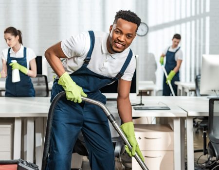 cheerful african american cleaner vacuuming floor while looking at camera